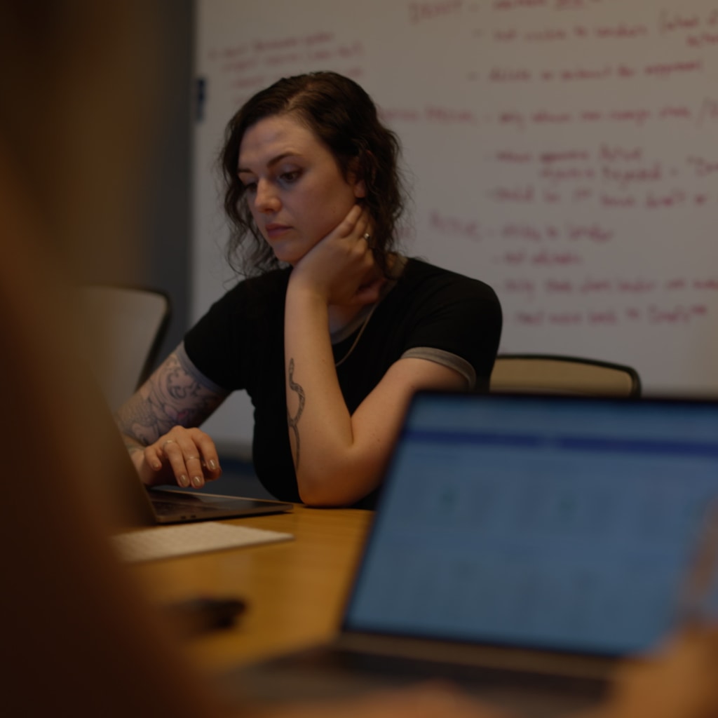 A software engineer at Cuttlesoft reviews generative AI model outputs, demonstrating our thoughtful approach to artificial intelligence implementation. Working with both laptop and external display, she analyzes model performance and fine-tuning results for custom AI solutions. The focused evaluation session, with code documentation visible on the whiteboard behind, shows how we carefully assess AI capabilities for enterprise applications. This detailed review process reflects Cuttlesoft's approach to generative AI - combining technical expertise with careful consideration of business impact and practical applications.